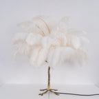 Front view of a Clérachic table lamp with unlit white ostrich feathers on a gold branch stem, with its black cord on a white surface.