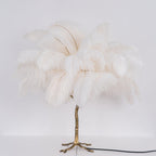 Lit Clérachic table lamp with white feathers glowing softly above a gold branch stem, with its black cord running across a white surface.