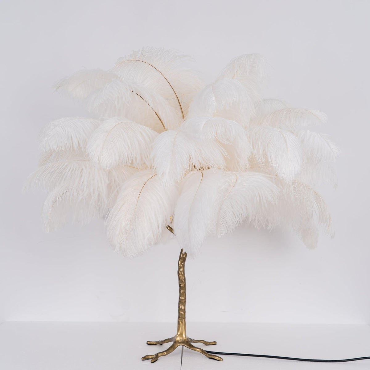 Lit Clérachic table lamp with white feathers glowing softly above a gold branch stem, with its black cord running across a white surface.