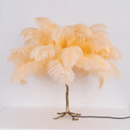 Clérachic table lamp with unlit peach-cream feathers on a gold branch stem and splayed feet, on a white surface.