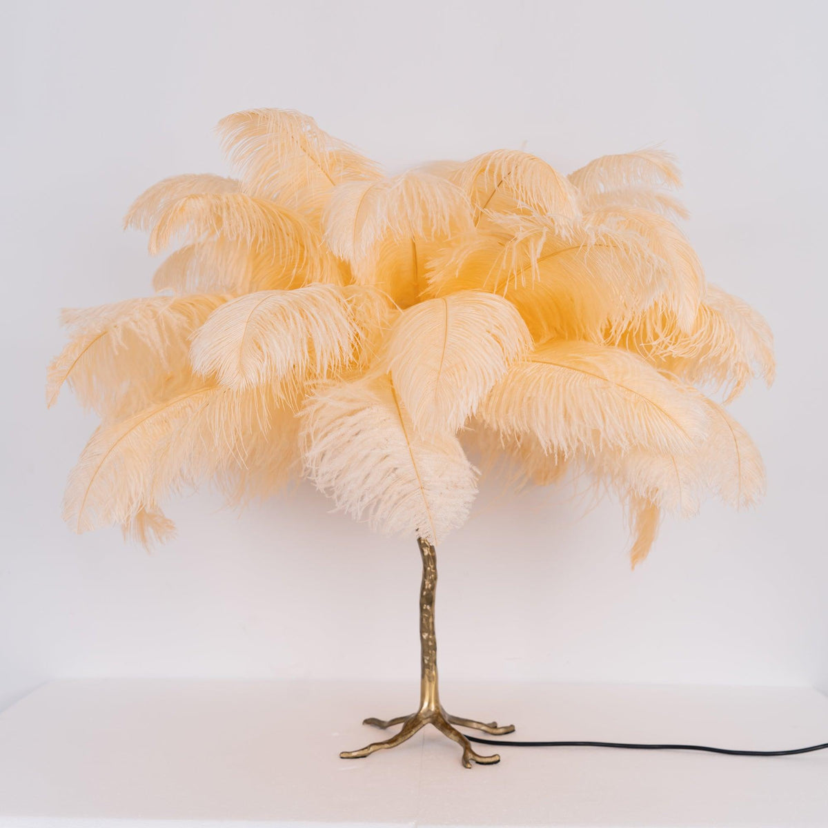 Clérachic table lamp with unlit peach-cream feathers on a gold branch stem and splayed feet, on a white surface.