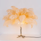 Lit Clérachic table lamp with peach feathers glowing amber on a gold branch stem, on a white surface with a black cord.