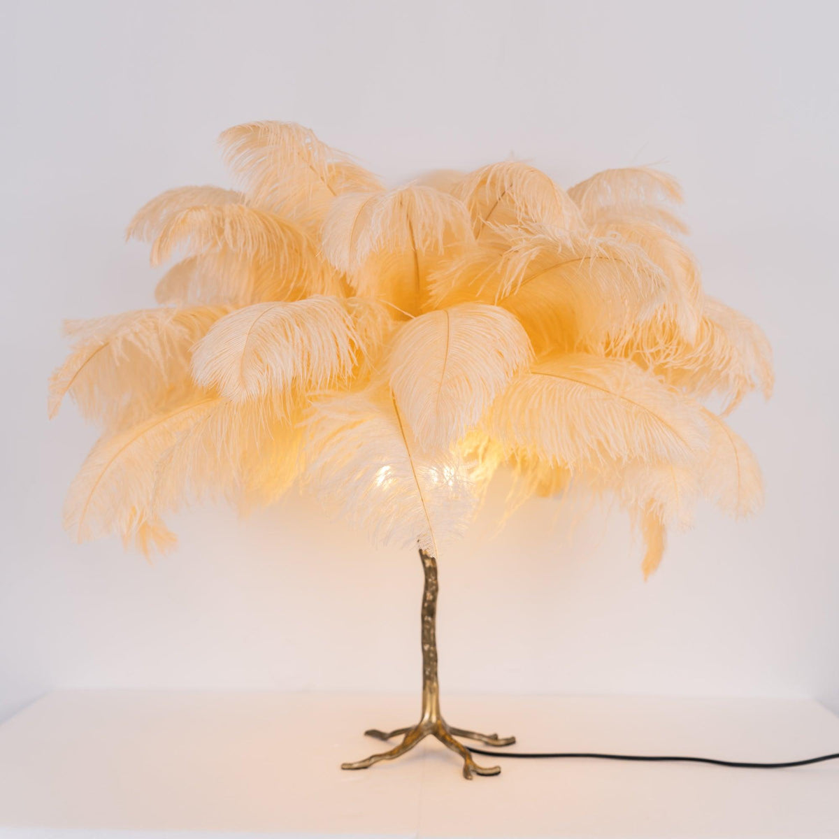 Lit Clérachic table lamp with peach feathers glowing amber on a gold branch stem, on a white surface with a black cord.