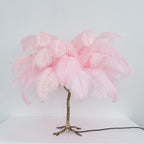 Clérachic table lamp with unlit candy-pink feathers on a gold branch stem, on a white surface in front of a pale blue wall.