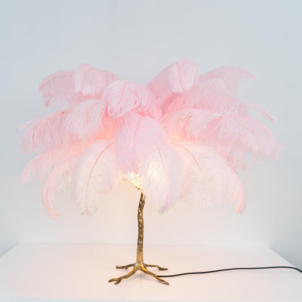 Lit Clérachic table lamp with pink feathers glowing at the centre above a gold branch stem, on a white surface.