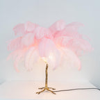 Lit Clérachic table lamp with pink feathers glowing at the centre above a gold branch stem, on a white surface.