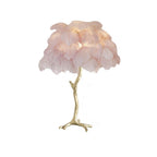 Clérachic table lamp with lit blush-pink feathers on a gold branch stem with splayed feet, on a white background.