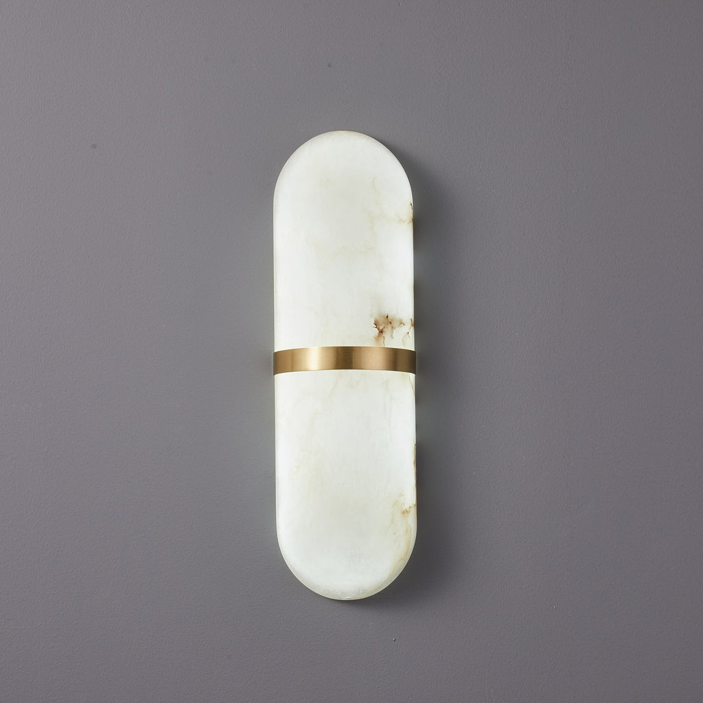 The Clérachic Ostruvyd pill sconce in white alabaster with a brushed brass band, mounted unlit on a soft grey-lilac wall.