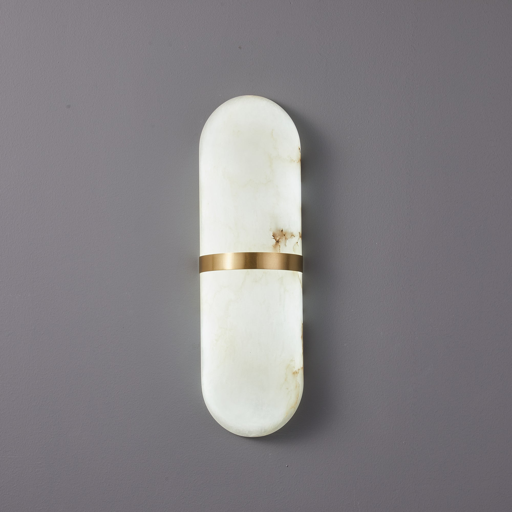 The Clérachic Ostruvyd pill sconce in white alabaster with a brushed brass band, mounted unlit on a soft grey-lilac wall.