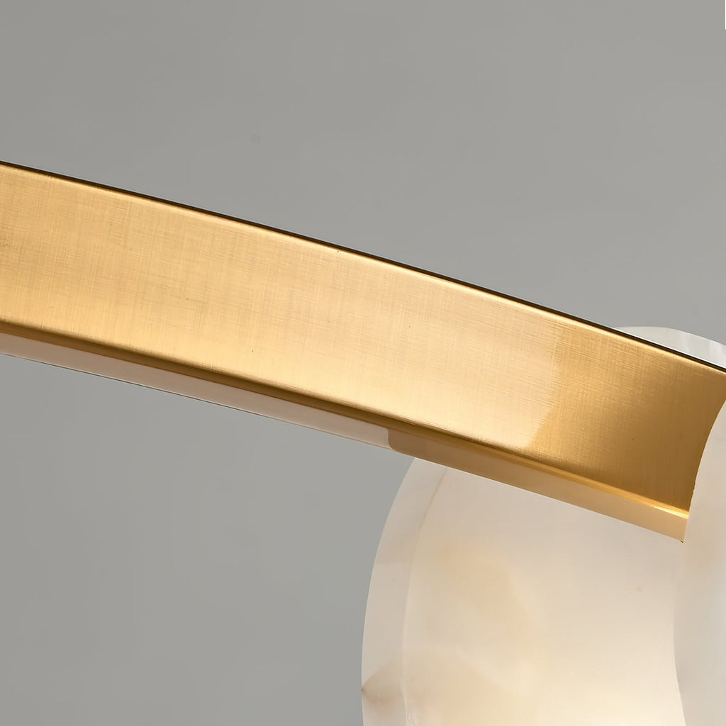 The Clérachic chandelier's brushed brass band close up, its satin grain curving past a translucent alabaster disc.