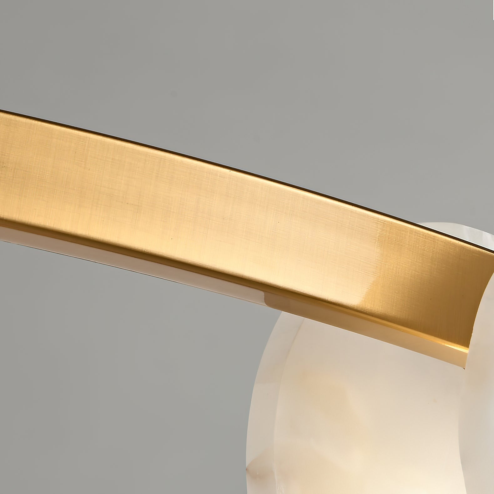 The Clérachic chandelier's brushed brass band close up, its satin grain curving past a translucent alabaster disc.