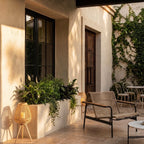 Clérachic tripod cane lanterns glowing on a sunlit terrace beside a planter, with a rope chair and bistro table behind.