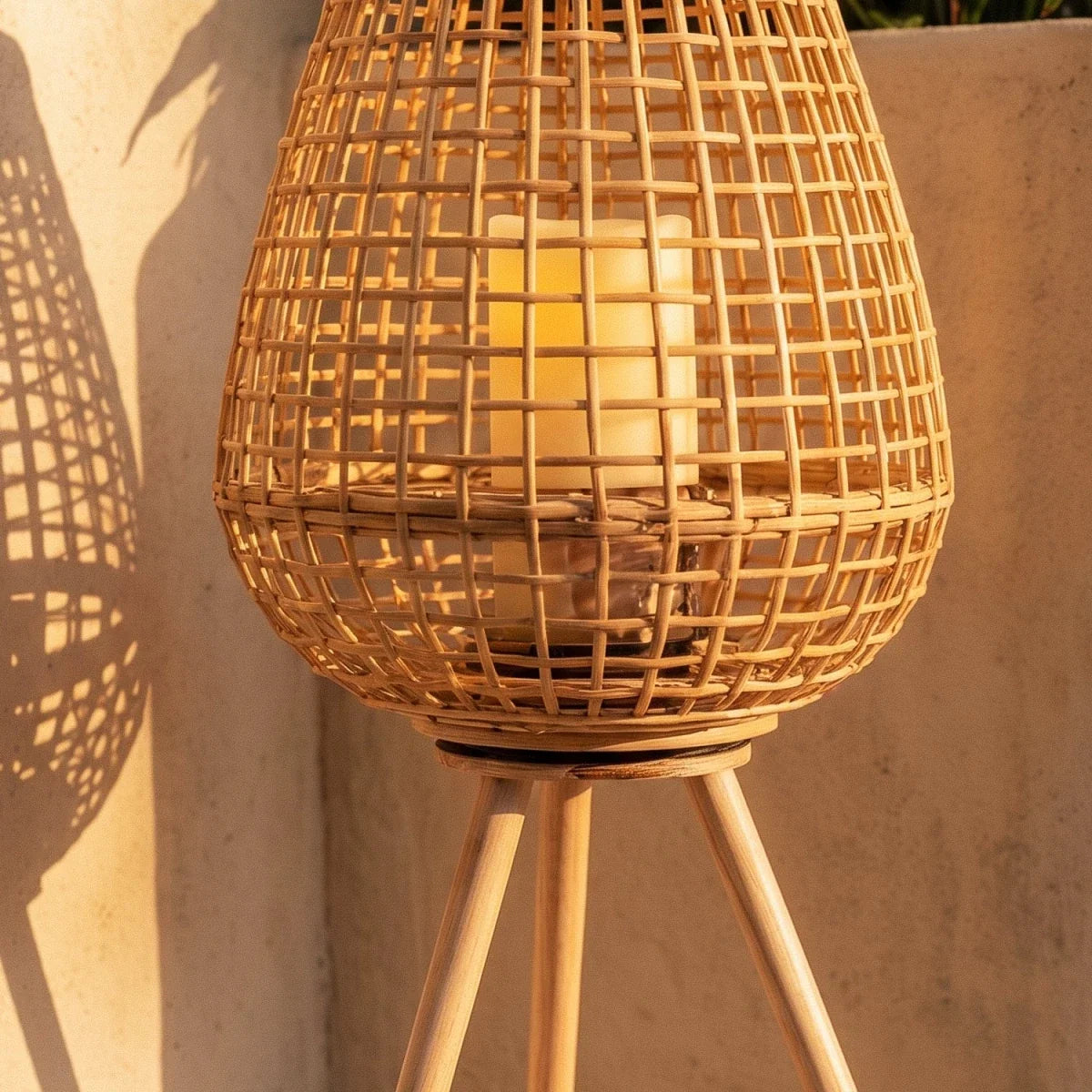 Clérachic close-up of the cane lantern on wooden tripod legs, a pillar candle glowing warm through the open weave.