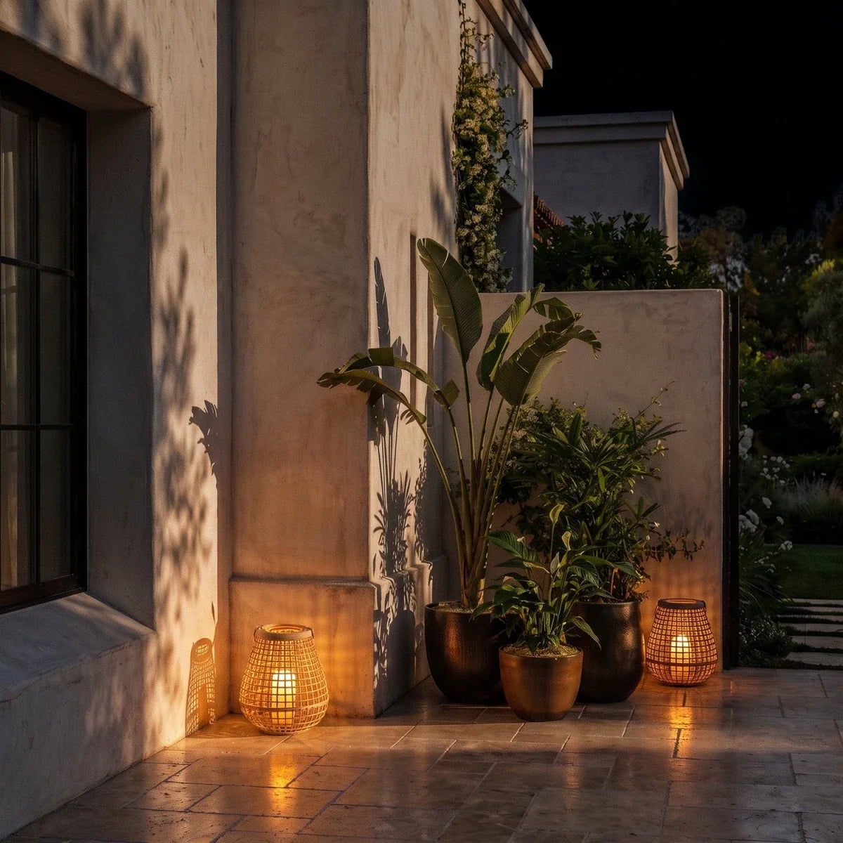 Clérachic two woven cane lanterns glowing on a terrace at night, beside terracotta planters and a plastered wall.