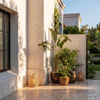 Clérachic woven cane lanterns on a sunlit paved terrace in the daytime, set among tall potted plants and a stucco wall.