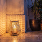 Clérachic woven cane lantern lit by a candle at dusk, casting a lattice of warm shadows over a stone terrace corner.
