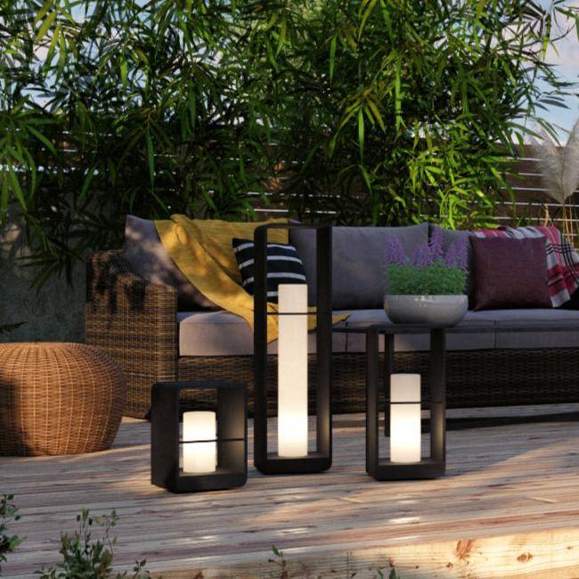 Clérachic three black lanterns in different heights on a timber terrace deck beside a rattan sofa, yellow throw and greenery.