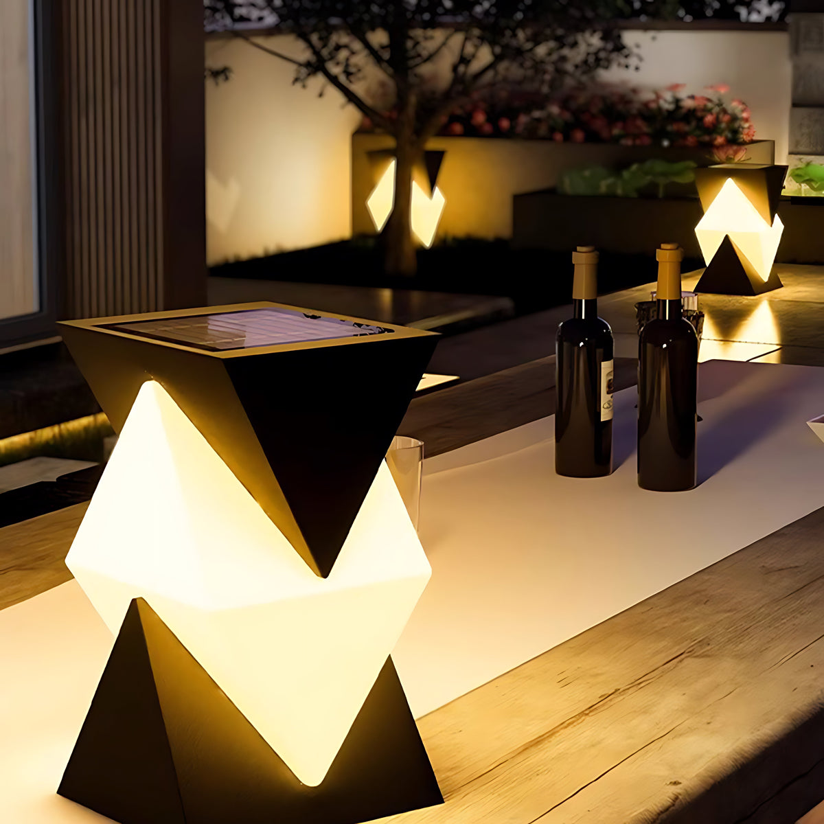 Clérachic solar outdoor pillar light glowing on an outdoor dining table beside two wine bottles, a garden wall blurred behind it.