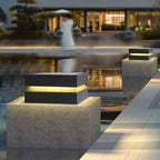 A Clérachic post light on a concrete block by still water, with a lit house reflected behind.