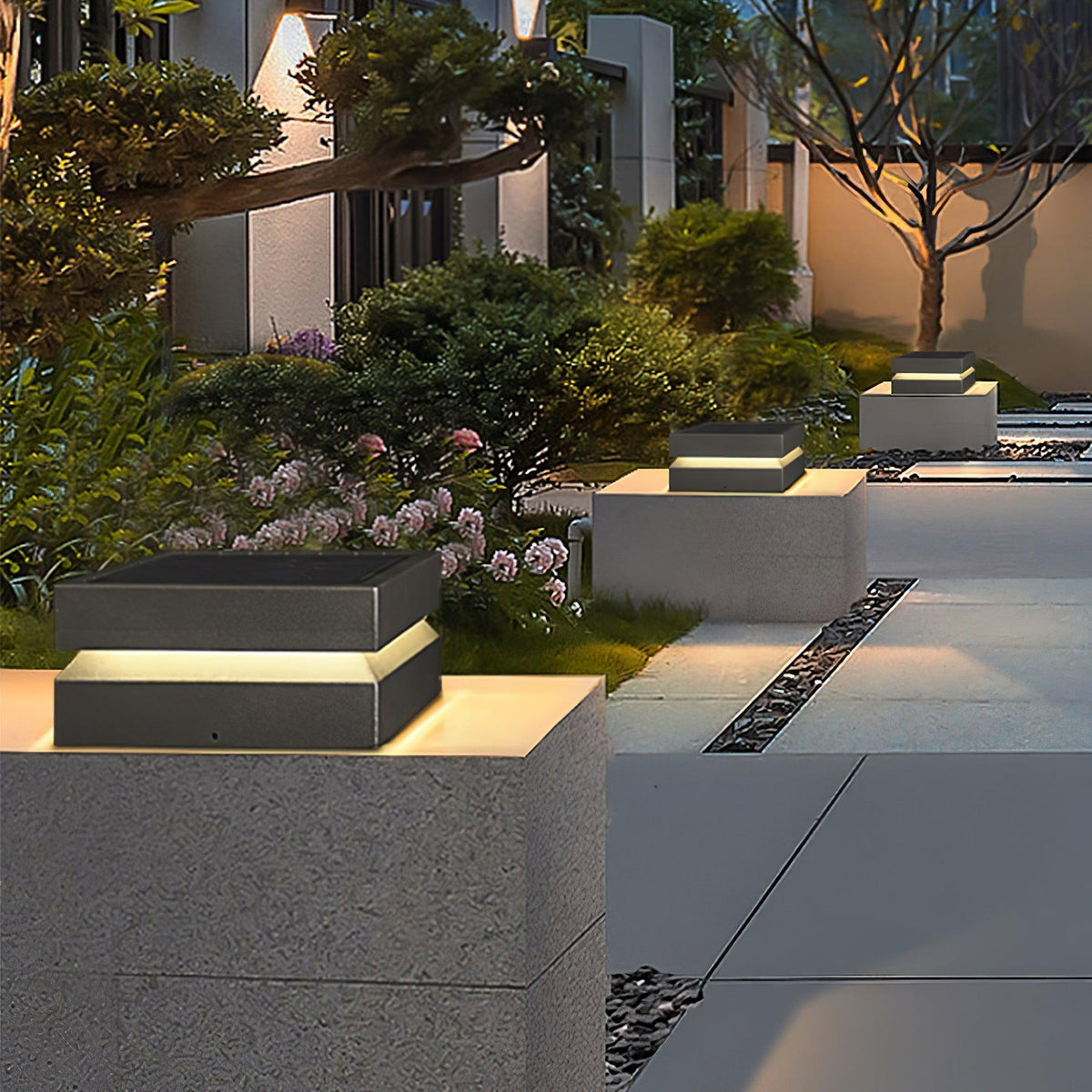 In a planted courtyard at dusk, a Clérachic post light sits on a pale block with more along the path.