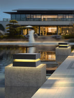 Across a reflecting pool, a Clérachic post light glows on a concrete block in front of a modern house.