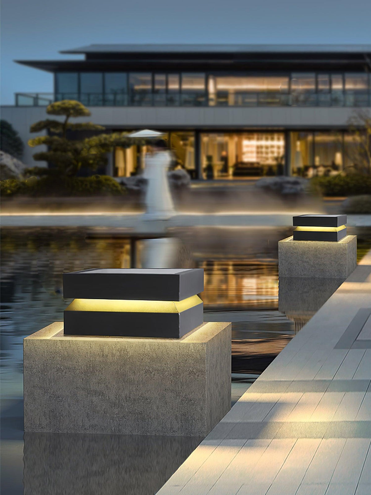 Across a reflecting pool, a Clérachic post light glows on a concrete block in front of a modern house.