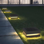 A line of Clérachic post lights marks a lawn edge beside a stone path, each glowing warm at night.