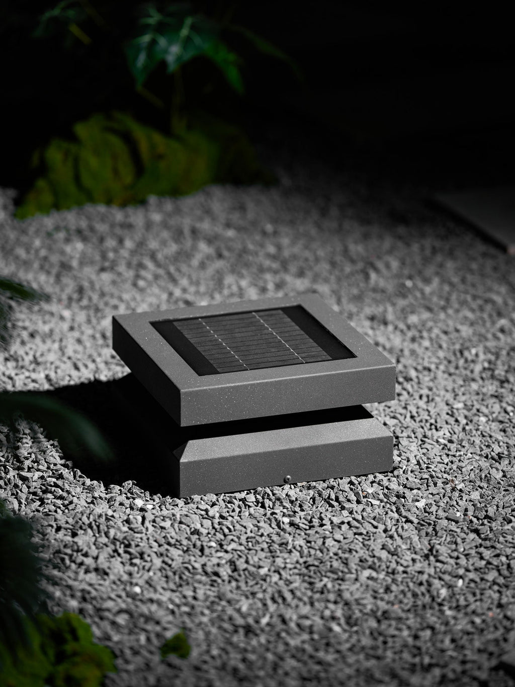 A Clérachic post light set low in grey gravel, unlit, with a green plant behind it.