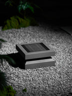 A Clérachic post light set low in grey gravel, unlit, with a green plant behind it.