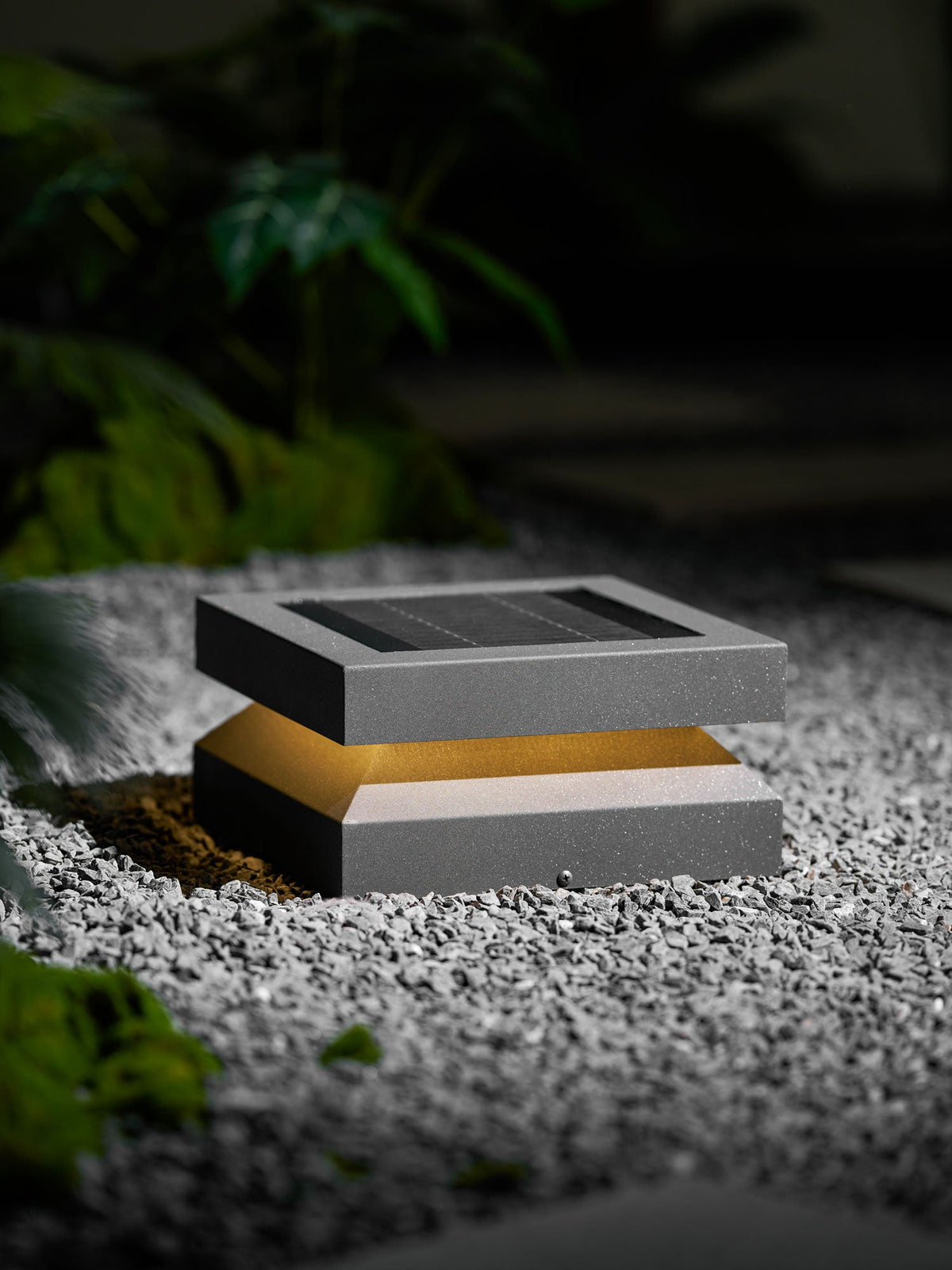 From a lower angle, a Clérachic post light on gravel glows softly beside a dark green plant.