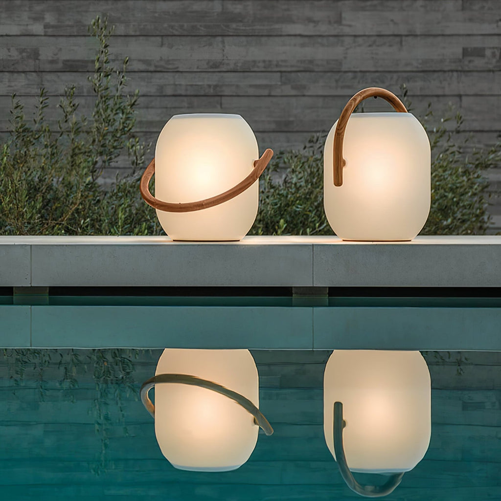 Two lit white Clérachic solar lanterns with wood-grain handles on a pool edge, their glow reflected in the still water.