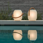 Two lit white Clérachic solar lanterns with wood-grain handles on a pool edge, their glow reflected in the still water.
