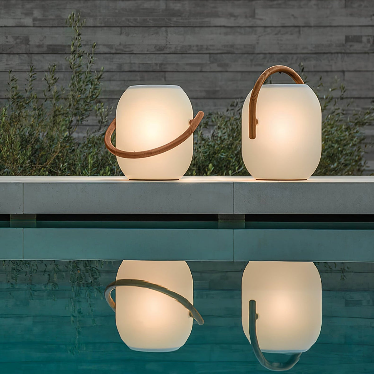 Two lit white Clérachic solar lanterns with wood-grain handles on a pool edge, their glow reflected in the still water.