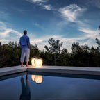 Two lit Clérachic lanterns on the edge of a pool at dusk, with a person in a blue shirt looking out over the trees.