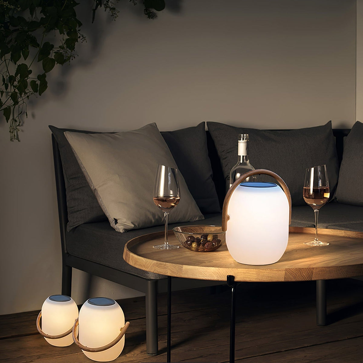 Clérachic solar lantern lit on a round wooden tray table between two wine glasses, with two more lanterns on the floor beside the sofa.
