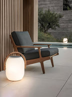 A Clérachic white lantern with a wood-grain handle glowing on a stone patio beside a teak lounge chair with dark cushions.