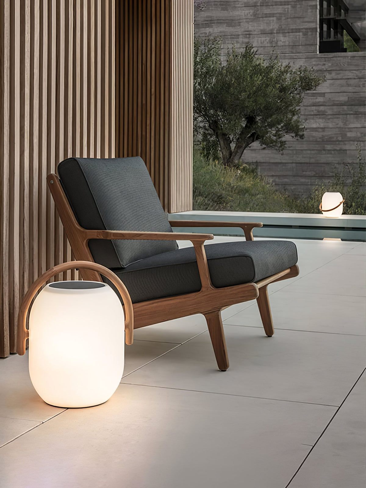 A Clérachic white lantern with a wood-grain handle glowing on a stone patio beside a teak lounge chair with dark cushions.