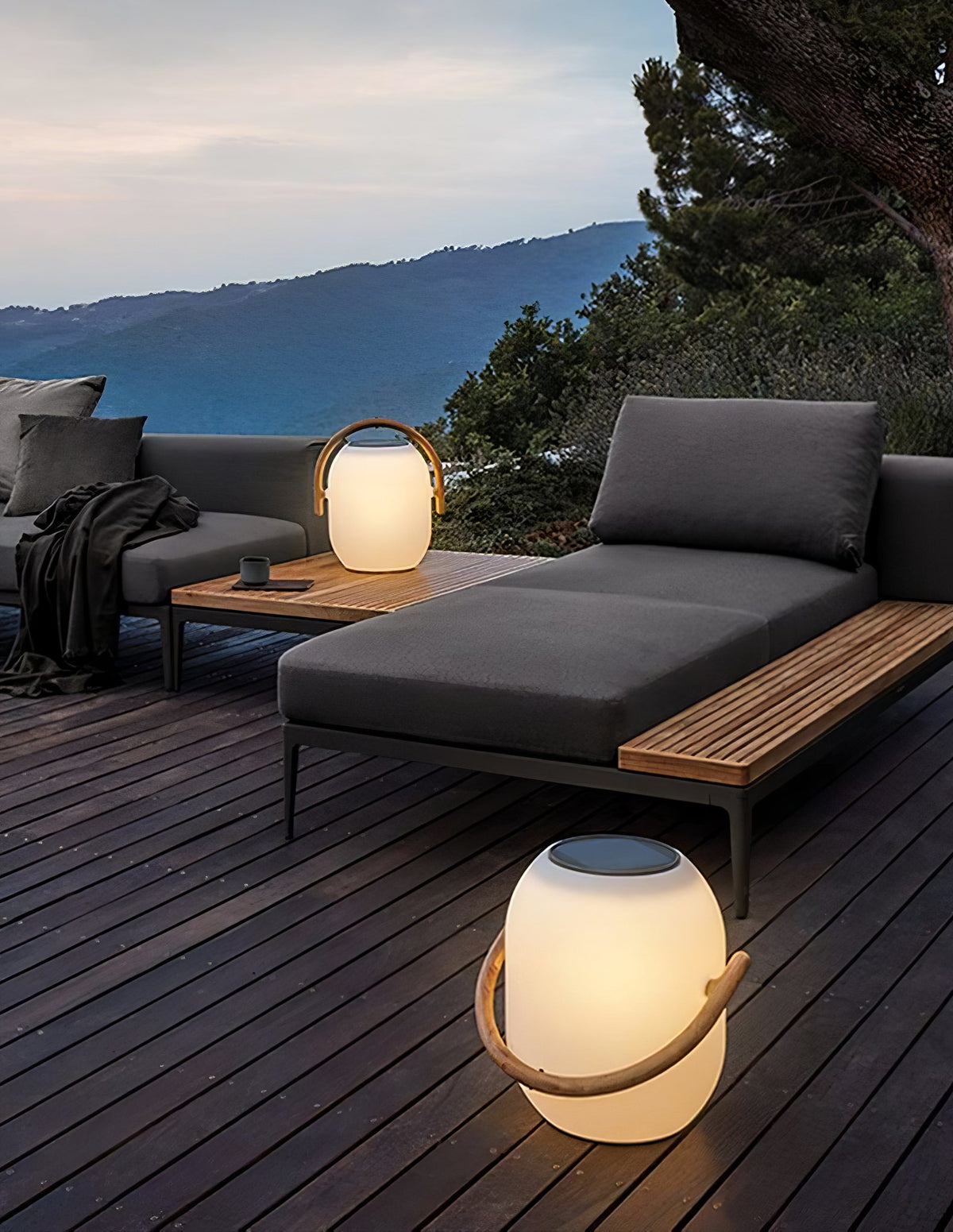 Lit Clérachic lanterns on a wooden deck at dusk, one on the boards and one on a low table beside a grey daybed.