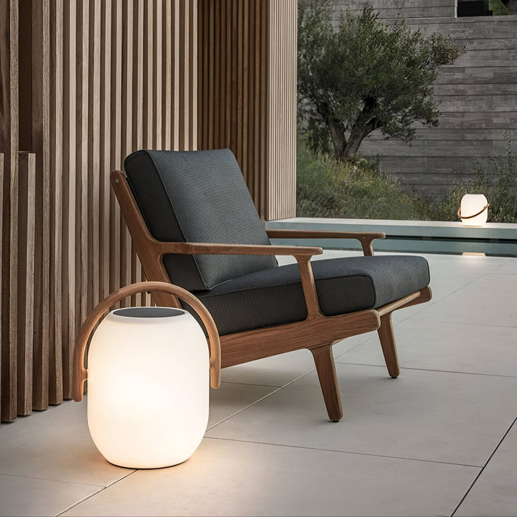 Close view of a lit Clérachic solar lantern on a pale terrace, in front of a wooden-framed armchair and slatted timber wall.