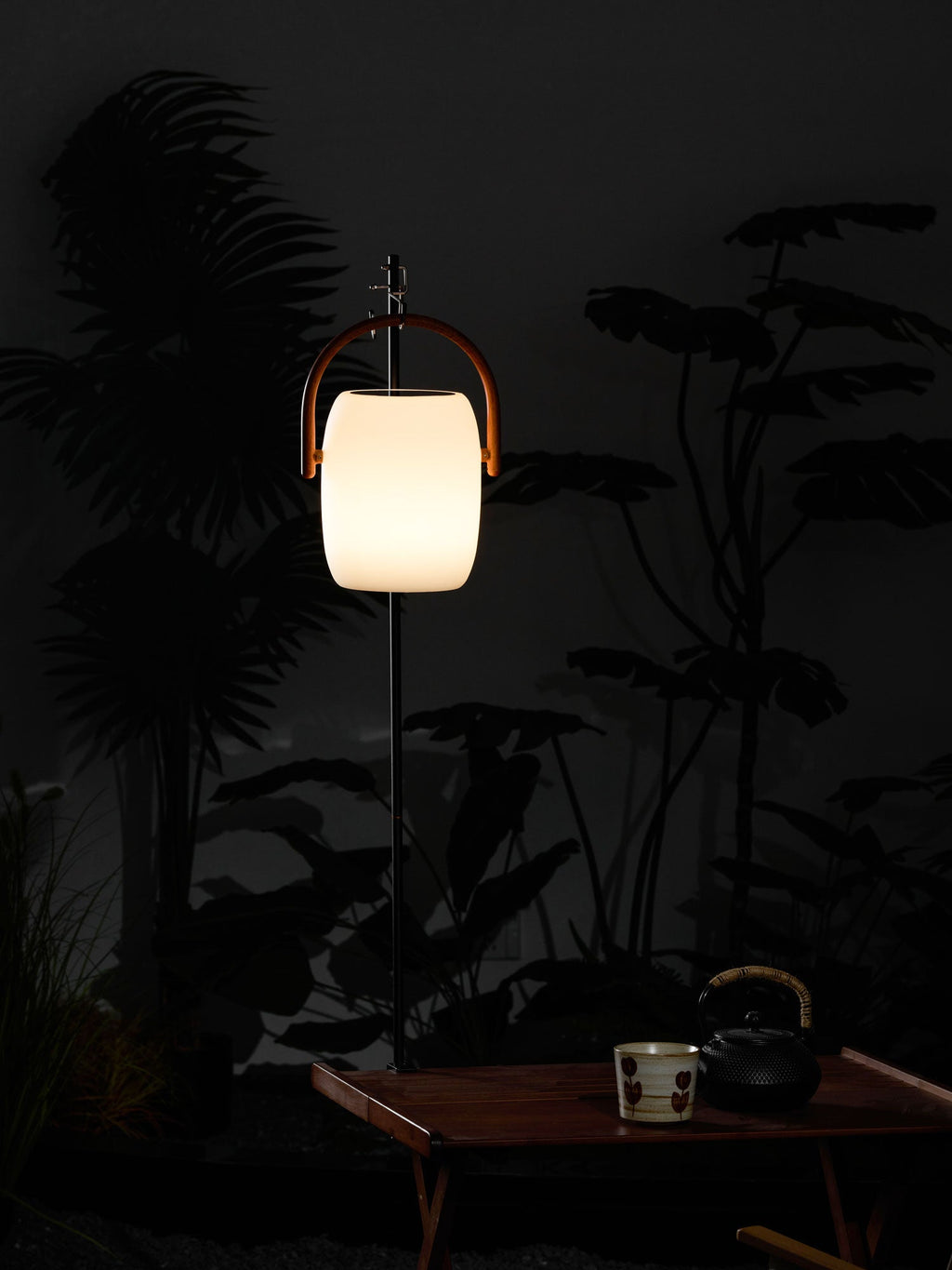 A Clérachic lantern hung from a slim pole beside dark leaves at night, lit warm, with a tea cup and kettle on a table below.