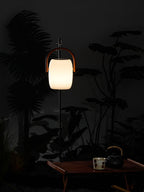 A Clérachic lantern hung from a slim pole beside dark leaves at night, lit warm, with a tea cup and kettle on a table below.