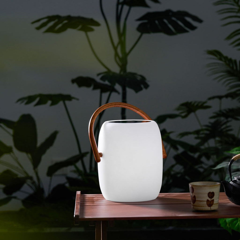 Unlit white Clérachic lantern with a wood-grain handle on a small wooden table among dark tropical leaves at night.