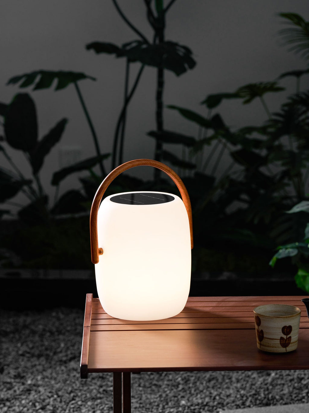 Softly lit Clérachic solar lantern on a slatted wooden table over gravel, a patterned cup beside it and leaves behind.