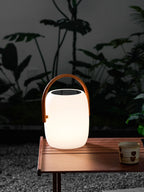 Softly lit Clérachic solar lantern on a slatted wooden table over gravel, a patterned cup beside it and leaves behind.