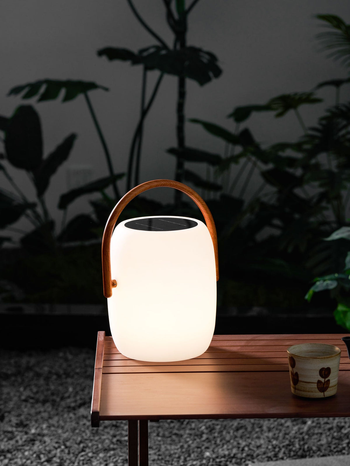 Softly lit Clérachic solar lantern on a slatted wooden table over gravel, a patterned cup beside it and leaves behind.