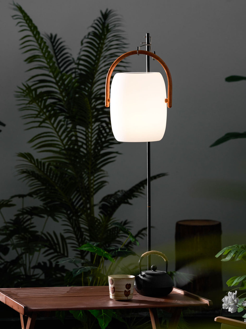 Clérachic lantern lit and hung from a black pole above a garden table, set against fern fronds and a dark wall.