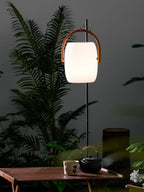 Clérachic lantern lit and hung from a black pole above a garden table, set against fern fronds and a dark wall.