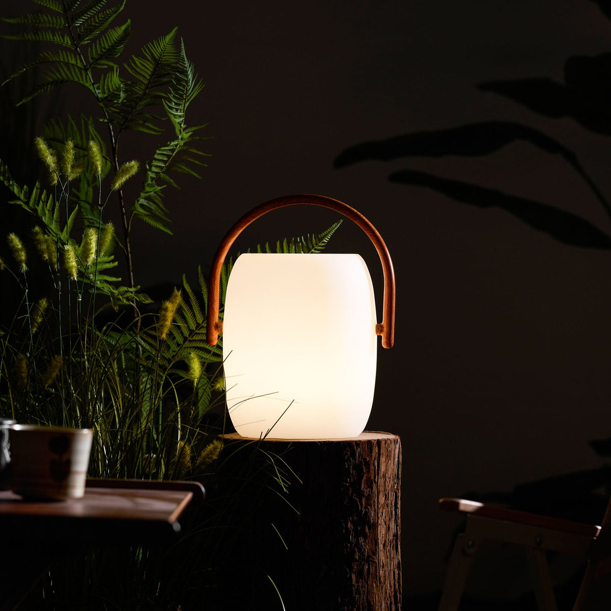 Clérachic solar lantern glowing warm on a tree stump, its wood-grain handle arched over it, grasses and ferns beside.