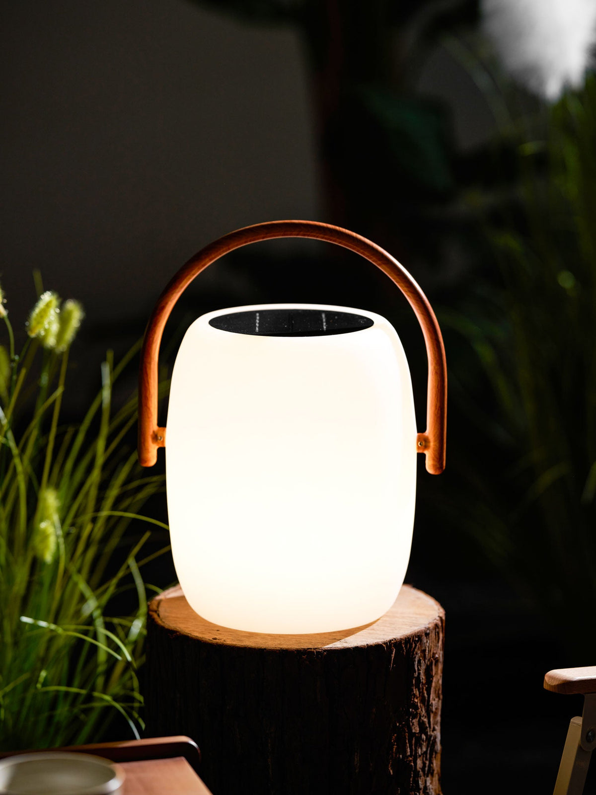 Evenly lit Clérachic lantern on a log slice, its dark solar panel on top and a wood-grain handle, in a dark garden.