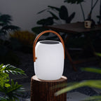 White Clérachic lantern, unlit, on a tree stump in a dim garden, with a wooden table and ferns around it.